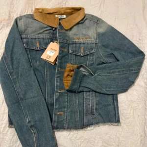 Jeansjacka från Miu Miu med manchesterkrage - Snygg jeansjacka från Miu Miu med ljusblå denim och kontrasterande beige manchesterkrage. Jackan har broderad logga på bröstet, bröstfickor med knappar och coola detaljer i brunt. Perfekt för dig som vill sticka ut med en unik touch. Passar mer xs