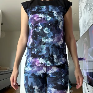 Blommig träningstopp i mesh + byxor (kan köpas separat) - Snygg träningstopp med blommigt mönster i blått, lila och svart. Toppen har meshpartier upptill och är ärmlös med rund hals. Perfekt för dig som vill sticka ut på gymmet eller löprundan. Har vuxen storlek S.    Kan köpas separat