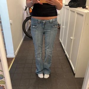 Lågmidjade ljusblå jeans med fickor - Säljer ett par lågmidjade ljusblå jeans med raka ben och snygga fickor framtill med lock och knapp. Jeansen har markerade sömmar längs benen och klassisk jeanslook. Perfekta för dig som gillar retrostil och vill ha något unikt i garderoben.