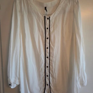 Vit långärmad skjorta från LYNX XL - Vit långärmad skjorta från LYNX i storlek XL. Skjortan har svarta knappar och svarta detaljer längs knappslån och över ryggen. Lös passform och rundad halsringning. Perfekt för dig som gillar stilrena och enkla plagg med en twist.