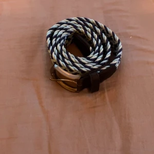 Randigt flätat bälte i blått och vitt - Snyggt flätat bälte med randigt mönster i marinblått och vitt. Spännet är i metall och detaljerna vid spännet är i mörkbrunt skinn. Perfekt accessoar för att lyfta din outfit med en sportig och fräsch känsla. Längd 110cm.