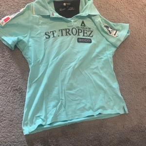 Turkos St. Tropez t-shirt med tryck - Turkos t-shirt från Les Voiles de St. Tropez med coolt tryck på bröstet och broderade patchar på ärmarna. Klassisk krage och korta ärmar, perfekt för sommarens häng. Skön bomullskvalitet och snygga detaljer som ger en sportig vibe.