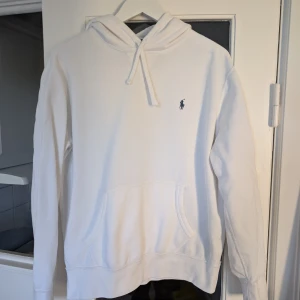 Vit hoodie från Polo Ralph Lauren - Snygg vit hoodie från Polo Ralph Lauren med klassisk broderad logga på bröstet. Tröjan har huva med snörning, känguruficka och långa ärmar. Tillverkad i mjuk bomull som är skön att ha på sig. Perfekt för en clean och stilren look.