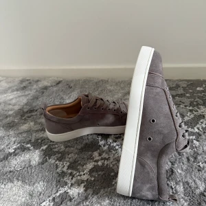 Grå mockasneakers från Louboutin - Snygga sneakers från Louboutin i grå mocka med klassisk röd sula och vita kanter. Skorna har rund tå, platt sula och snörning. Perfekta för dig som gillar stilrena och exklusiva detaljer.