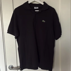 Svart pikétröja från Lacoste - Klassisk svart pikétröja från Lacoste med den ikoniska gröna krokodilloggan på bröstet. Tröjan har korta ärmar, krage och knäppning framtill. Perfekt för en clean och sportig look. Tillverkad i mjuk bomull som känns skön mot huden.