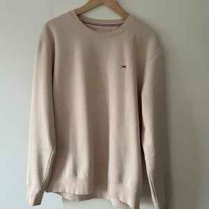 Beige sweatshirt från Tommy Hilfiger XL - Snygg beige sweatshirt från Tommy Hilfiger i storlek XL. Tröjan har rund hals, ribbade muddar och ett litet broderat logomärke på bröstet. Tillverkad i mjuk bomullsmix som ger en clean och modern vibe. Perfekt att matcha med jeans eller chinos.