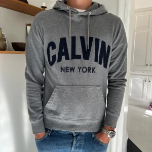 Grå hoodie från Calvin Klein - Snygg grå hoodie från Calvin Klein med stor svart text 'CALVIN NEW YORK' på bröstet. Tröjan har huva med snörning, magficka och långa ärmar. Mjuk bomullskvalitet och normal passform.