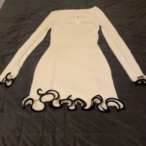 Vit kort klänning med volangkant - Supersnygg vit klänning från Bik Bok med lång ärm och coola svarta volangdetaljer vid ärmslut och nederkant. Klänningen är figurnära och har en modern, stilren look som sticker ut med sina vågiga kanter. Perfekt för dig som gillar trendiga detaljer.