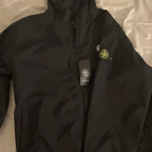 Svart vindjacka från Stone Island - Svart vindjacka från Stone Island i storlek XL. Jackan har klassisk logga på ärmen, huva och dragkedja framtill. Snygg och stilren modell i lätt material, perfekt för dig som gillar streetwear och vill ha något som funkar året runt.