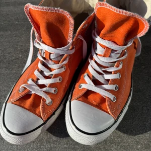 Orange Converse All Star high tops - Säljer ett par orangea Converse All Star high tops med klassisk vit gummitå och vita snören. Skorna har rund tå, platt sula och det ikoniska All Star-märket på sidan. Ovandelen är i slitstark bomullstextil och sulan i gummi. Perfekt för dig som gillar färgstarka sneakers.