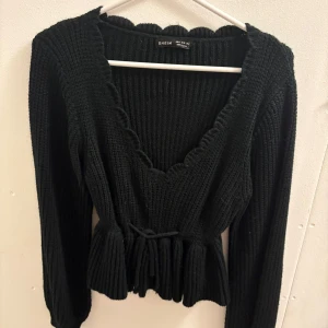 Svart stickad kofta från SHEIN - Supersöt svart stickad kofta från SHEIN med scallop-kantad v-ringning och knyt i midjan. Modellen är croppad med volangdetalj nedtill och har långa ärmar. Perfekt för dig som gillar trendiga och mysiga plagg med lite extra detaljer. 