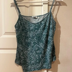 Grön mönstrad linnetopp från Indiska - Snygg grön/ blå/ turkos  linnetopp från Indiska i storlek M med tunna axelband och ett detaljerat paisley-inspirerat mönster i vitt. Toppen har en avslappnad passform och är perfekt för varma dagar. Materialet känns lätt och luftigt mot huden. Storlek M men jag brukar ha s/xs och den passar bra på mig 