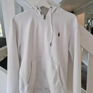 Vit hoodie från Polo Ralph Lauren - Säljer en stilren vit hoodie från Polo Ralph Lauren med dragkedja framtill och klassisk logga på bröstet. Tröjan har huva med snörning, två fickor och är långärmad. Perfekt för chill dagar och enkel att matcha med jeans eller joggers.