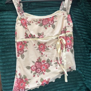 Blommig linnetopp med rosett - Supersöt linnetopp i ljusbeige med rosa och röda blommor, gröna blad och en söt rosett i satinband vid midjan. Toppen har breda axelband och vågig nederkant för extra detalj. Perfekt för en somrig vibe!