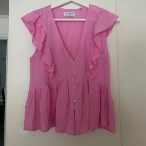 Rosa volangblus från Holly & Whyte - Supersöt rosa blus med volanger längs axlarna och fram, v-ringning och knappar hela vägen ner. Luftig och flowy modell som ger en chill och feminin vibe. Perfekt för dig som gillar färg och detaljer!💗 Aldrig använd