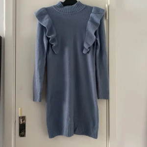 Blå stickad klänning med volang - Supersöt ljusblå stickad klänning med långa ärmar och volangdetaljer på axlarna. Hög ribbad hals och ribbade muddar ger en mysig känsla. Perfekt för dig som gillar en feminin och trendig look.