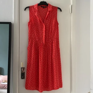 Röd prickig ärmlös kortklänning med fickor - Superfin röd ärmlös klänning med vita prickar och krage. Klänningen har knappar framtill och ett lätt, luftigt tyg som faller snyggt. Perfekt för varma dagar när du vill känna dig både söt och bekväm.