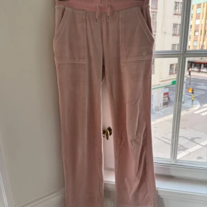 Rosa mjukisbyxor Juicy Couture XS - Supermjuka ljusrosa mjukisbyxor från Juicy Couture med bred resår och snörning i midjan. Byxorna har raka ben, stora fickor fram och bakfickor med knapp. Tillverkade i glansig polyesterblandning för den klassiska Juicy-looken.