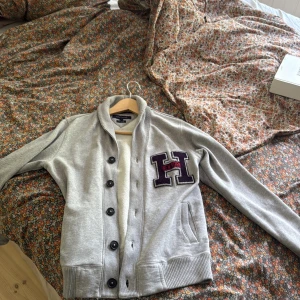 Grå kofta med H New York patch - Snygg grå kofta från Tommy Hilfiger med stor H New York patch i lila och svart på bröstet. Koftan har svarta knappar, ribbade muddar och två fickor framtill. Perfekt för dig som gillar college-stil och vill ha något bekvämt och stilrent.