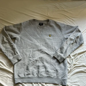 Grå sweatshirt från Lyle & Scott - Grå sweatshirt från Lyle & Scott Junior med klassisk rund hals och långärmade muddar. På bröstet sitter den ikoniska gula fågeln broderad. Tröjan är gjord i mjuk bomull och har en clean, enkel look som funkar till jeans eller joggers. Storlek Junior 14/15 år pris kan diskuteras 