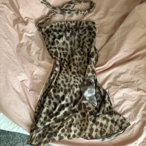 Leopardmönstrad klänning - Säljer en kort leopardmönstrad klänning  med volangdetaljer och axelbandslös design. Klänningen har bruna och beige toner och är tillverkad i ett lätt, transparent material som ger en cool och trendig vibe.