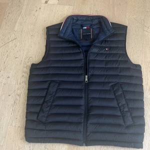 Svart dunväst Tommy Hilfiger L - Svart dunväst från Tommy Hilfiger i storlek L. Västen har horisontella quiltade paneler, två sidofickor och dragkedja framtill. Hög krage med diskret röd, vit och blå detalj på insidan. Klassisk och stilren design med liten logga på bröstet. Säljer den pga att den är för stor