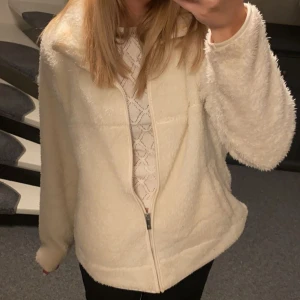 Beige teddyjacka med dragkedja - Mysig beige teddyjacka med hel dragkedja framtill och hög krage. Jackan har en fluffig och mjuk känsla, perfekt för kyliga dagar. Passformen är normal och modellen är enkel utan synliga detaljer eller mönster.