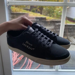Svarta Saint Laurent sneakers - Svarta sneakers från Saint Laurent med vit logga broderad på sidan. Skorna har svarta snören, rund tå och vit platt sula. Ovandelen är i canvas och detaljer i skinn, vilket ger en clean och stilren vibe. Perfekta för dig som gillar minimalistisk och lyxig streetstyle.
