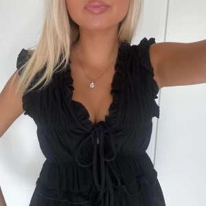 Svart volangblus  - Supersöt svart blus med volangdetaljer och knyt framtill. Djup v-ringning och ärmlös design med rynkade partier💓💗