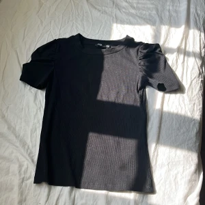Svart ribbad topp med puffärm - Svart ribbad topp från NewYorker i storlek XXS. Modellen har korta puffärmar som ger en snygg siluett och rund halsringning. Materialet är mjukt och stretchigt, perfekt för dig som gillar en enkel men trendig look.