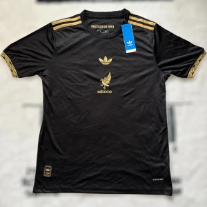 Svart och guld Mexiko fotbollströja Adidas - Snygg svart fotbollströja från Adidas med guldiga detaljer och broderade loggor. Tröjan har korta ärmar med guldfärgade mönster på mudden och tre klassiska ränder på axlarna. Framsidan pryds av Adidas-logga och Mexikos örn i guld. Tillverkad i lätt och ventilerande material.
