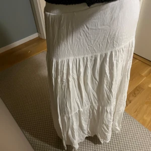 Vit långkjol i bomull - Säljer en flowy vit långkjol med volangdetaljer. Kjolen har en lätt skrynklig struktur och är perfekt för dig som gillar en avslappnad och bohemisk stil. Den är hög i midjan och har en luftig känsla, vilket gör den superbekväm att bära.