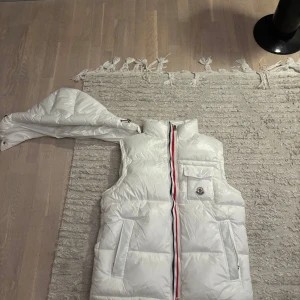 Vit dunväst från Moncler - Snygg vit dunväst från Moncler med hög krage och klassisk Moncler-logga på bröstfickan. Västen har en dragkedja med röd, vit och blå rand, två sidofickor och en bröstficka med lock. Materialet är glansigt och quiltat, perfekt för kyliga dagar.