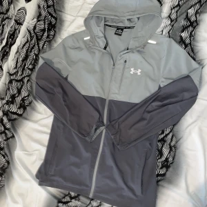 Grå och svart vindjacka från Under Armour - Snygg tvåfärgad vindjacka från Under Armour i grått och svart. Jackan har huva, hel dragkedja och diskret logga på bröstet. Perfekt för blåsiga dagar och träning utomhus. Lätt och smidig design med långa ärmar och praktiska fickor.