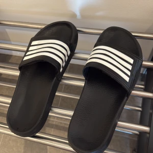 Svarta Adidas sandaler med vita ränder - Storlek 42 ungefär, passar inomhus och utomhus