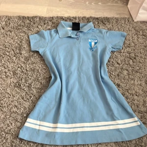 Ljusblå kortklänning med krage från lag mff - Sportig ljusblå kortklänning med klassisk pikékrage och korta ärmar. Klänningen har två vita ränder längst ner och ett tryckt emblem på bröstet. Materialet är mjuk bomull, perfekt för en chill och aktiv stil.