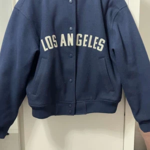 Blå bomberjacka H&M Divided - Mörkblå bomberjacka från H&M Divided med 'LOS ANGELES' tryckt i vitt på bröstet. Jackan har ribbade muddar, knappar framtill och två snedställda fickor. Perfekt för dig som gillar sportig och avslappnad stil.