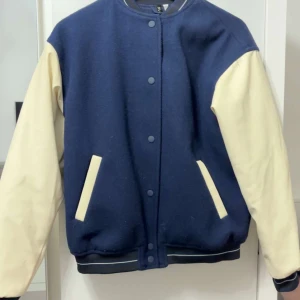 Blå och beige bomberjacka H&M Divided - Snygg bomberjacka från H&M Divided i blått med beige ärmar. Jackan har tryckknappar framtill, ribbade muddar och två fickor med beige detaljer. Perfekt för dig som vill ha en klassisk varsity-look med modern twist.