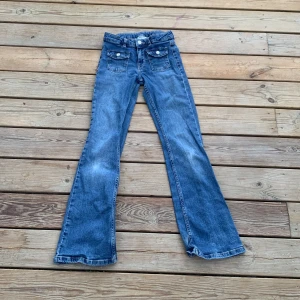 Blå bootcut jeans från Kappahl - Snygga blå bootcut jeans från Kappahl med blekning på knäna. Jeansen har framfickor med lock och knappdetalj. 