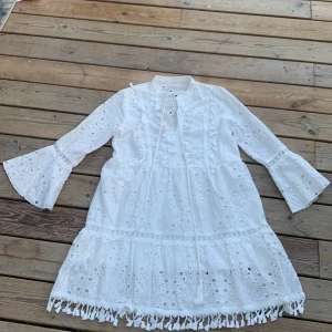 Vit student klänning  - Superfin vit kortklänning med broderade detaljer och hålmönster. Klänningen har trumpetärmar, knytband vid halsen och dekorativa fransar längst ner. Den är köpt i Italien och har inte kommit till användning en ända gång.