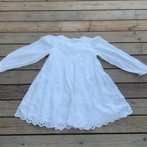 Vit broderad studentklänning från Zara - Superfin vit klänning från Zara med långärmade puffärmar och broderade blommönster över hela plagget. Klänningen har en kort, utsvängd passform och rund halsringning. 