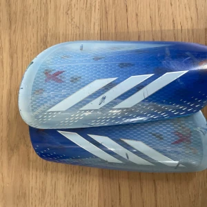 Adidas benskydd X blå/vit - Tja, säljer mina Adidas X benskydd. Det medkommer ett par övertäckande strumpor för att fästa benskydden bättre runt benet. Benskydden är i bra, men använt skick.