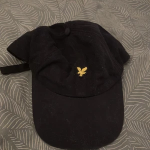 Svart manchesterkeps från Lyle & Scott - Snygg svart keps i manchester från Lyle & Scott med klassisk gul broderad örn framtill. Justerbar rem baktill för perfekt passform. Perfekt accessoar för en cool och avslappnad stil.