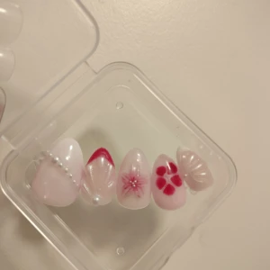 Press-on naglar med pärlor och blommor - Snygga handgjorda press-on naglar i rosa och rött med olika dekorationer som pärlor, 3D-blommor och glansig finish. Perfekt för dig som vill ha unika naglar snabbt och billigt. 10 at, kommer i en genomskinlig ask och en bubble wrap påse. Finns i Storlek S, M, L. Skriv gärna privat om det finns någon annan design som ni skulle vilja ha, så fixar jag det!
