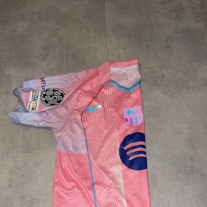 FC Barcelona rosa matchtröja Nike - Säljer en FC Barcelona fotbollströja från Nike i rosa och ljuslila med blå detaljer. Tröjan har korta ärmar, Dri-FIT-material och tryckta loggor från både Nike och Spotify. Snygg design med tunna linjer och klubbmärke på bröstet.