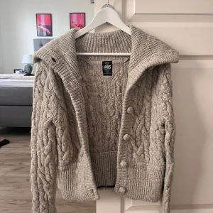 Grå kabelstickad kofta med krage - Säljer en grå kabelstickad kofta från Tommy Hilfiger med bred ribbad krage och knappar framtill. Koftan har lång ärm, grovstickat mönster och breda muddar. Perfekt för dig som gillar mysiga och stilrena plagg med lite retrovibe.