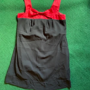 Svart och röd klänning/top Vero Moda - Supersöt kort klänning/top från Vero Moda i svart med röd överdel och breda axelband. Snygg rosettdetalj framtill. Perfekt för dig som vill sticka ut med en färgklick. Materialet känns lätt och skönt mot huden.