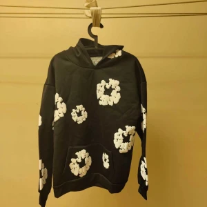 Denim Tears - Svart hoodie från Denim Tears med unika vita blomliknande tryck över hela tröjan. Klassisk huva, magficka och lång ärm. Perfekt statement-plagg för dig som vill sticka ut med streetwear-vibe.