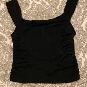 Svart topp från SHEIN - Snygg svart topp från SHEIN i mjukt smidigt tyg och breda axelband. Modellen är croppad och har lätt rynkade detaljer på sidorna för en cool look. Perfekt för dig som gillar trendiga och stilrena plagg.