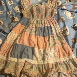 Patchwork pastellklänning Lindex Kids - Supergullig kortklänning från Lindex Kids i patchworkstil med pastelliga färger som ljusblå, rosa, beige och vitt. Klänningen har volangkant, spetsdetaljer och korta ärmar. Materialet är mjukt bomullstyg med blommigt och mönstrat tyg i olika sektioner.